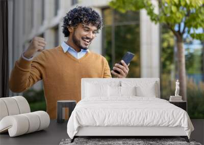 Smiling young man in casual attire celebrating success with raised fist and smartphone in hand. Energetic expression shows happiness, achievement, and positivity. Wall mural