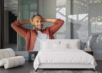 Smiling young man relaxing in office with laptop. Concept of success, happiness, relaxation. Man in casual attire reflecting achievement in modern workspace. Wall mural