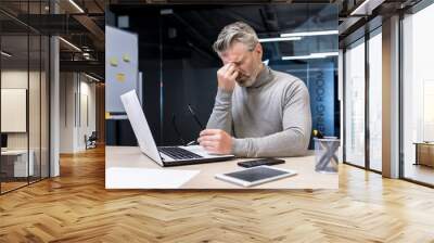 Stressful, tense, tired handsome male businessman sitting in office at desk with laptop and phone. Takes a break, took off his glasses, covered his eyes with his hand. Wall mural