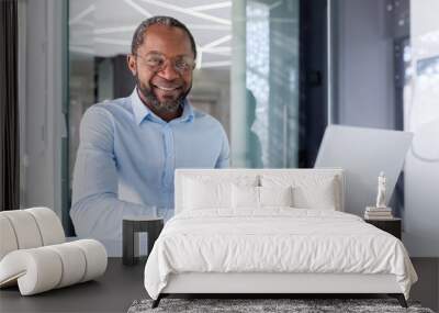 Successful african american businessman in shirt and glasses working inside bright modern office, man smiling and looking at camera, portrait of successful boss using laptop sitting at workplace. Wall mural