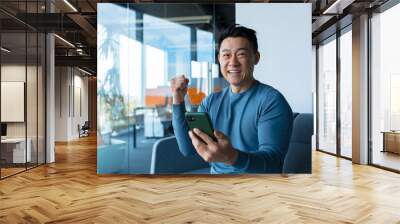 Successful and happy asian businessman man celebrating victory sitting and working in modern office at desk, celebrating victory looking at camera and joyfully shouting holding mobile phone Wall mural