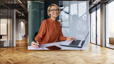 Successful and happy business woman looking at camera sitting inside office working with documents, portrait of satisfied woman writing in notebook and working using laptop at work. Wall mural