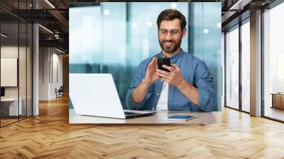 Successful and happy businessman with beard works in modern bright office, male boss uses laptop, business owner in casual shirt holds phone, browses internet pages Wall mural