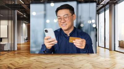 Successful and smiling asian businessman in office making bank transfer and online purchase in online store, man using app on phone and bank credit card, close up inside. Wall mural