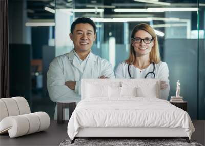 Successful and smiling doctors look at the camera and smile, with folded arms, working in a modern clinic, an Asian man and a female team of colleagues Wall mural