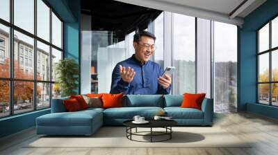 Successful asian boss near window received good news online, businessman use phone and celebrate victory and good achievement results inside office, man in shirt and glasses excitedly rejoice. Wall mural