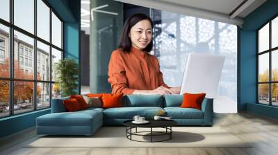 Successful asian business woman working inside office building with laptop, female worker smiling and looking at computer screen, satisfied with work achievement results. Wall mural