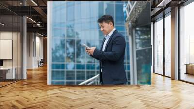 Successful Asian businessman in business suit walking outside near office building, male manager director using phone, smiling and happy reading online message. Wall mural