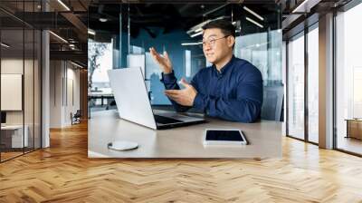 Successful Asian businessman talking on video call using laptop for remote communication with colleagues, boss in shirt smiling and gesturing with hands, online meeting with partners. Wall mural