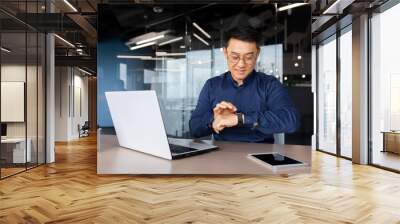 Successful asian businessman use smart watch at workplace, man chooses apps and smile happy with apps, employee inside office use laptop at work. Wall mural