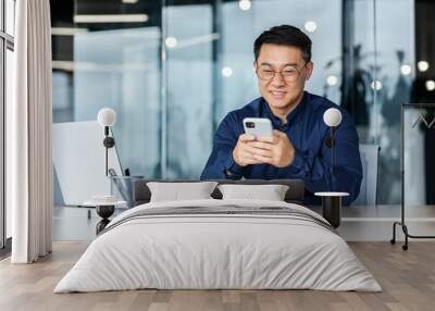 Successful Asian businessman working inside modern office building, man smiling and happy using smartphone to browse online pages, man in shirt and glasses with laptop. Wall mural