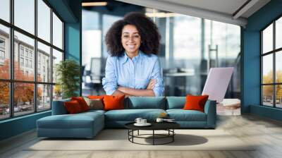 Successful black woman in blue shirt leaning with folded hands on table with laptop and daybook. Smiling lady looking at camera during working day as secretary in leading financial company. Wall mural