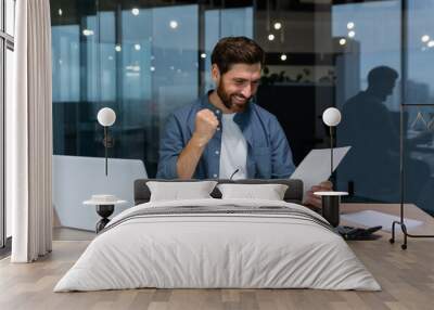 Successful businessman in casual shirt doing paperwork, boss with beard and glasses sitting at desk at workplace using laptop working with documents, holding hand up celebrating victory and triumph. Wall mural