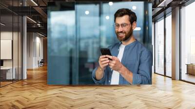 Successful businessman in glasses and beard standing near window in the evening, man using phone, happy boss typing message and browsing online pages on smartphone app inside office. Wall mural