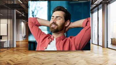 Successful businessman in office in a red shirt rests with his hands folded head with closed eyes, mature man with a beard works with a laptop at the workplace inside, dreams of future achievements. Wall mural