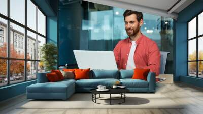 Successful businessman in red shirt happily working with laptop inside office, mature man with beard at workplace typing on keyboard smiling satisfied with work results and achievement. Wall mural