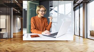 Successful businesswoman inside the office is satisfied with the results of the achievement, the employee received a good completed technical task, holds her hands up in a gesture of triumph Wall mural