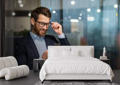 Successful financier investor works inside office at work, businessman in business suit uses telephone near window, man smiles and reads good news online from smartphone. Wall mural