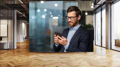 Successful financier investor works inside office at work, businessman in business suit uses telephone near window, man smiles and reads good news online from smartphone. Wall mural