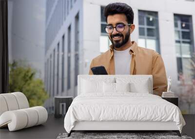 Successful Indian young businessman outside office building walking in daytime, man holding phone in hands, businessman dialing, browsing online pages, programmer engineer in glasses. Wall mural