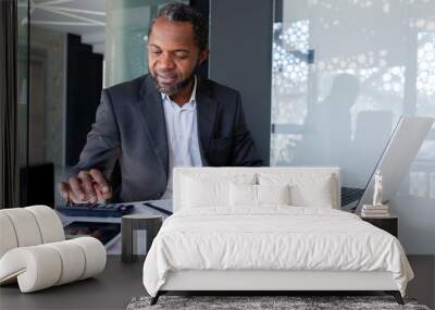 Successful satisfied financier behind paper work, afro american senior experienced filling out documents, using calculator for calculation, businessman behind paper work inside office smiling. Wall mural