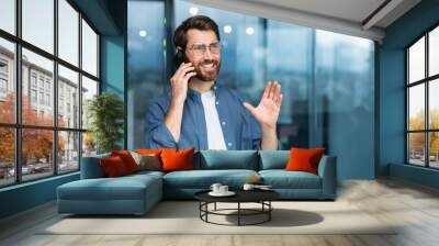 Successful smiling businessman in casual shirt talking on phone, male boss in glasses and beard near window inside modern office. Wall mural