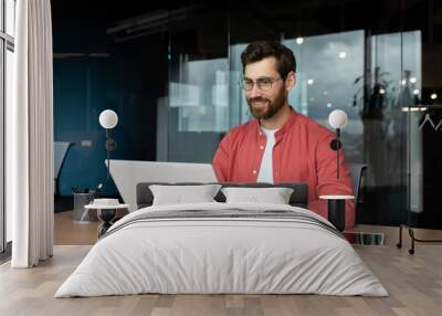 Successful smiling man working inside office with laptop, businessman in red shirt smiling and typing on keyboard in glasses, programmer working software for program. Wall mural