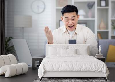 The winner is sitting at the table at home, the man is happy to receive the online notification of the victory and success on the phone, the Asian man is holding his hand up and holding smartphone. Wall mural