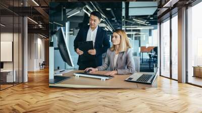 Two Asian male and female colleagues in a modern office, a woman shows the work done on the monitor, consults and discusses Wall mural