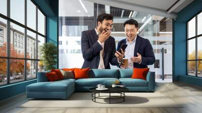 Two colleagues sharing a light moment in office while watching a funny video on smartphone. Both men dressed in business suits, one holding a coffee cup, expressing laughter and surprise. Wall mural