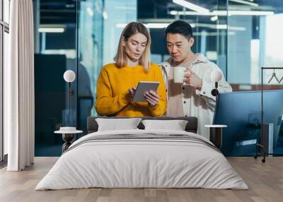 Two employees are having fun. during the break an Asian man and a blonde woman, colleagues work together in a modern office, look at a tablet computer together Wall mural