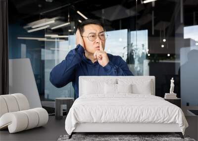 Upset and nervous asian man at workplace demanding silence from colleagues, businessman holding finger near lips and quarreling with co-workers, row inside office at workplace, discomfort at work. Wall mural