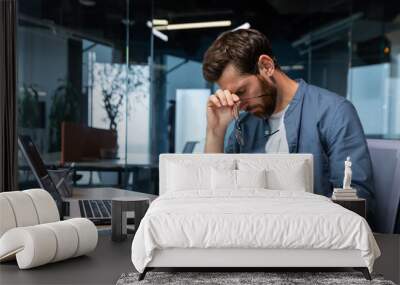 Upset businessman behind paper work inside modern office, mature man with beard reading financial reports and account documents unhappy with results and disappointed with achievements. Wall mural