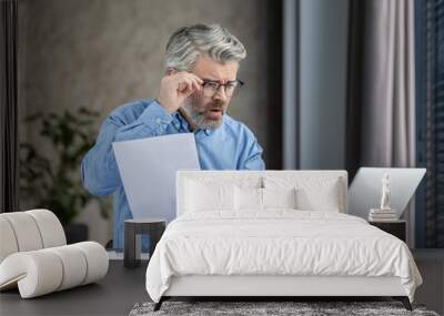 Upset businessman reading a document in his office, feeling concerned about its contents. Working on a laptop. Professional and office environment. Wall mural