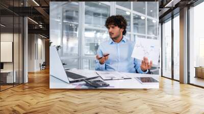 Upset financier showing online report, man working inside office nervously showing document, hispanic man in online meeting using laptop for remote communication and video call. Wall mural