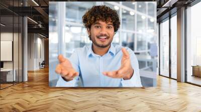 Video call online conference meeting, young successful hispanic man talking to colleagues remotely sitting in modern office, man looking at camera and smiling talking to colleagues in blue shirt. Wall mural