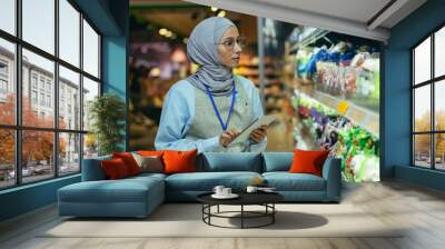 woman Seller in super market in hijab with tablet checking products using pocket computer, Muslim woman near shelves with products and goods. Wall mural