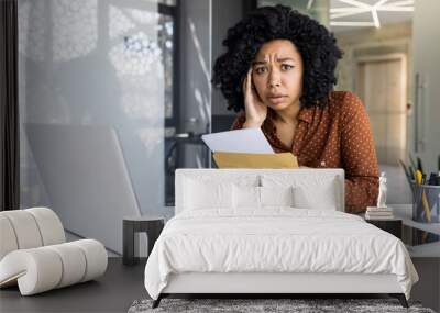 Young African American woman looks concerned while reading a letter at her modern office desk, surrounded by laptop and documents. Wall mural