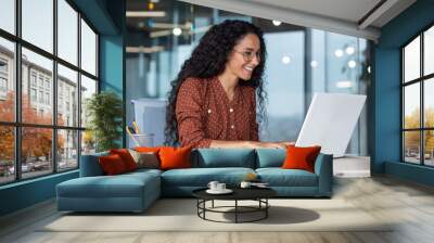 Young and happy hispanic woman working in modern office using laptop, business woman smiling and happy in glasses and curly hair. Wall mural