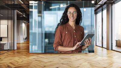 Young beautiful and successful business woman with tablet computer and glasses, smiling and looking out the window, Hispanic woman working inside a modern office building at work Wall mural