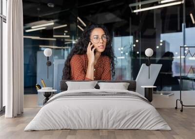 Young beautiful businesswoman talking on the phone and looking towards the window, happy hispanic woman inside the office using a laptop at work Wall mural