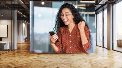 Young beautiful hispanic woman with curly hair inside office celebrating victory success, businesswoman received win notification on phone, using smartphone app at workplace Wall mural