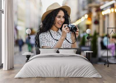 Young beautiful hispanic woman with curly hair walking in the evening city with a camera, female tourist on a trip exploring historical landmarks in the city. Wall mural