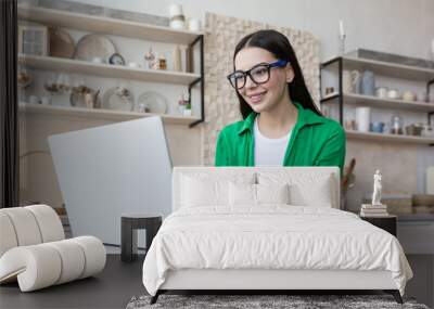 Young beautiful woman at home in the kitchen in glasses and a green shirt makes online purchases in an online store, brunette uses a laptop and holds a bank credit card in her hand Wall mural