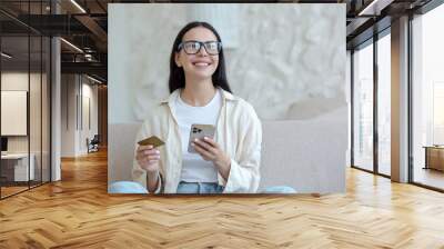 Young beautiful woman in glasses smiling and happy doing online shopping in online store sitting on sofa at home, holding mobile smartphone and bank credit card Wall mural