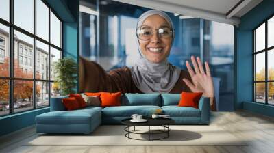 Young beautiful woman in hijab looking at smartphone camera, talking with friends on video call, muslim woman in glasses smiling and waving greeting gesture, working inside office modern building Wall mural