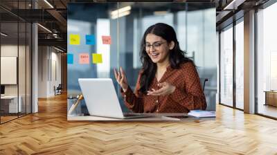 Young business woman inside office using laptop for video call, female worker smiling and talking with colleagues remotely in online meeting, joyful female worker with computer. Wall mural