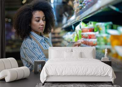 Young female grocery store worker using a tablet to manage inventory in an aisle filled with various products. Wall mural