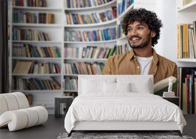 Young hispanic student reading a book while standing inside an academic university library, man smiling contentedly looking towards the window. Wall mural