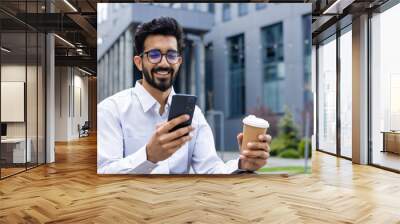 Young man with phone in hands and a cup of hot drink close up, businessman smiling contentedly browsing social networks, worker resting outside office building, using smartphone. Wall mural